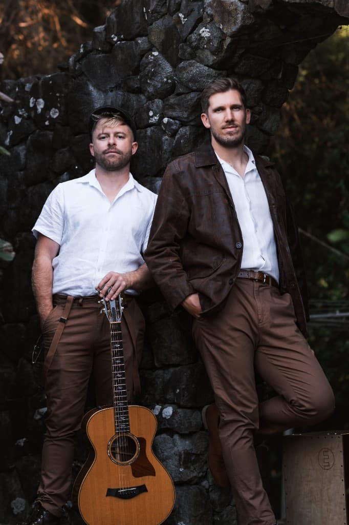 Professional portrait of a two-man band posing with an acoustic guitar in front of a rustic stone wall