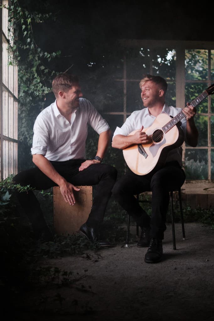 Sam and James performing together in an ivy-lined sunroom with guitar and cajon